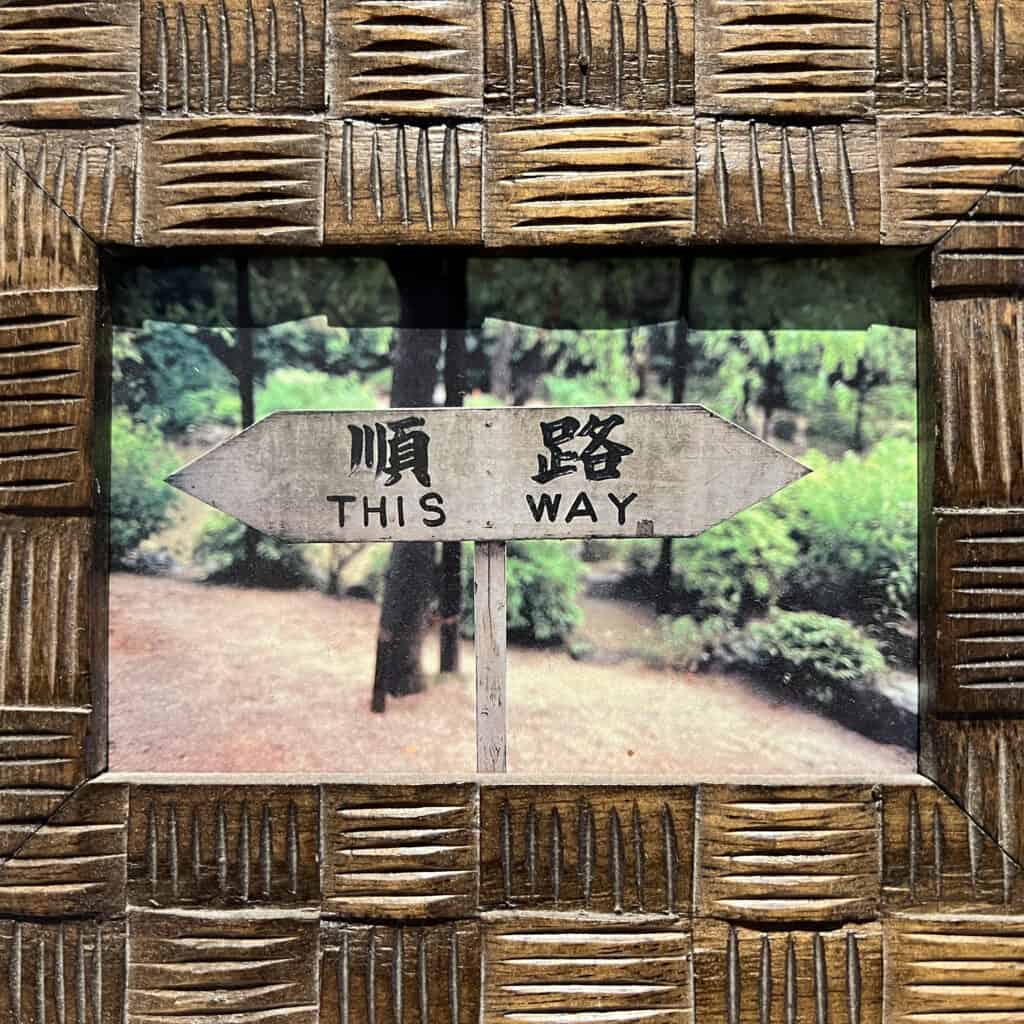 A tactile earthy wooden frame with a photo in it with a "This Way" sign written in Chinese and English, in the forest with a trail in the background